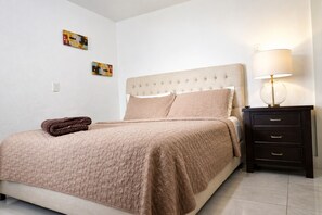 Un lit bien fait, recouvert d’une couette beige, une table de chevet en bois foncé avec une lampe, et deux tableaux abstraits accrochés au mur.