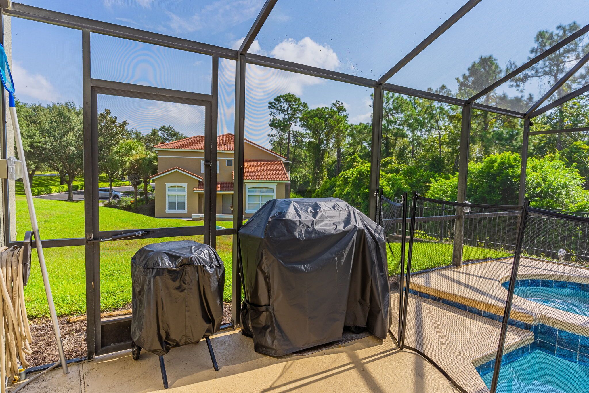 A screened-in area with a view of a house, a swimming pool, and covered barbecue grills.