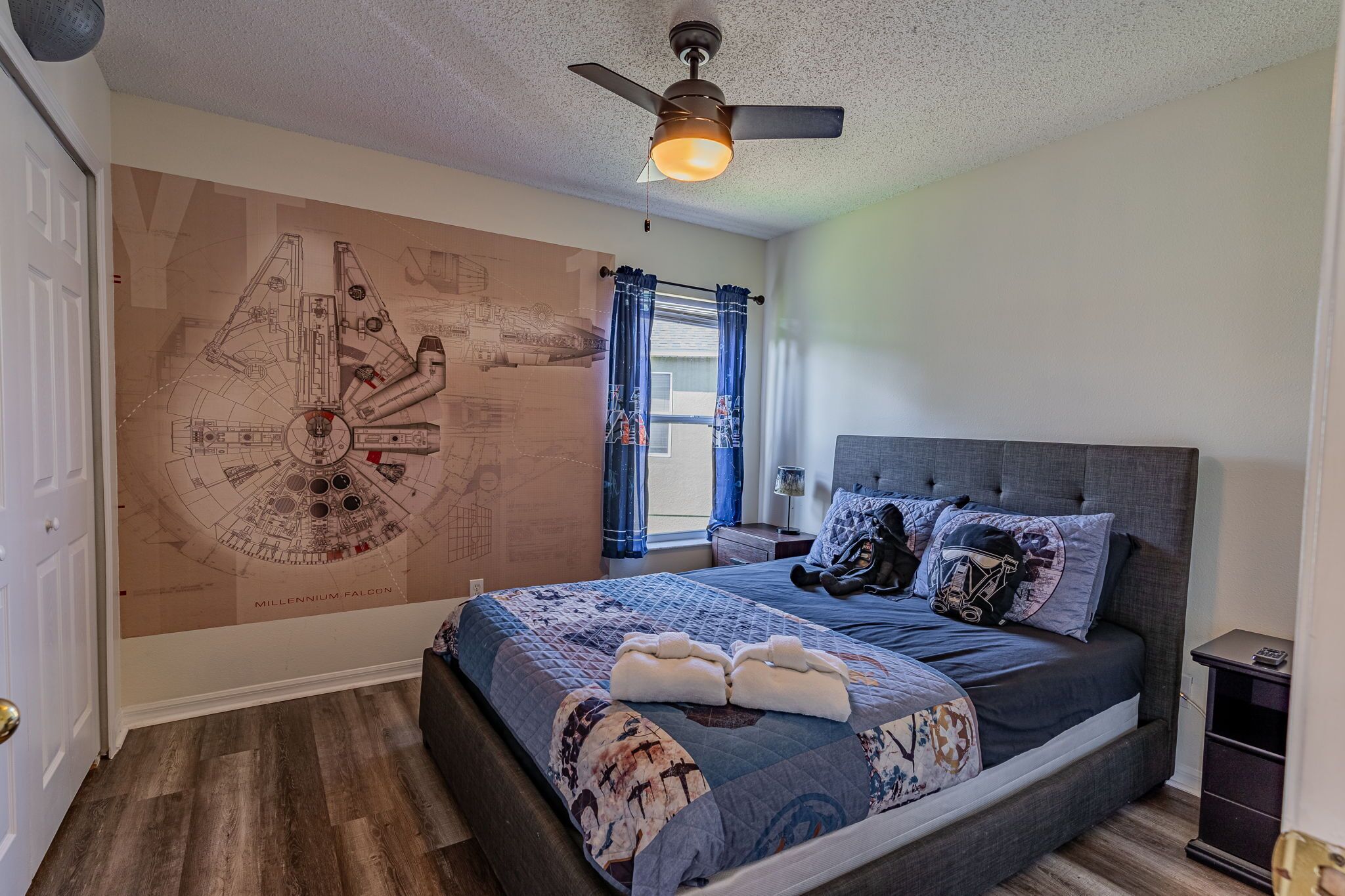 A bedroom with a large wall mural of a spaceship, a bed with a blue quilt, and a ceiling fan.