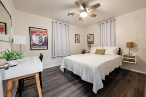 Un dormitorio con cama, escritorio, ventilador de techo y ventana con cortinas.