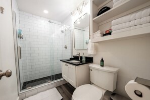 Un baño moderno con ducha de vidrio, paredes de azulejos blancos, lavamanos con encimera negra y un inodoro blanco.