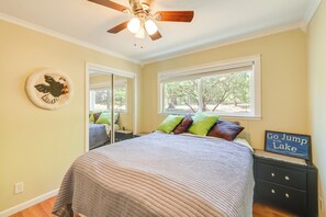 Une chambre avec un lit, un ventilateur de plafond, une fenêtre donnant sur des arbres, un miroir et un panneau sur la commode.