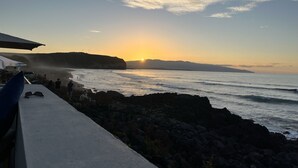 Een kustgezicht met een ondergaande zon, een rotsachtige kustlijn en mensen op het strand.