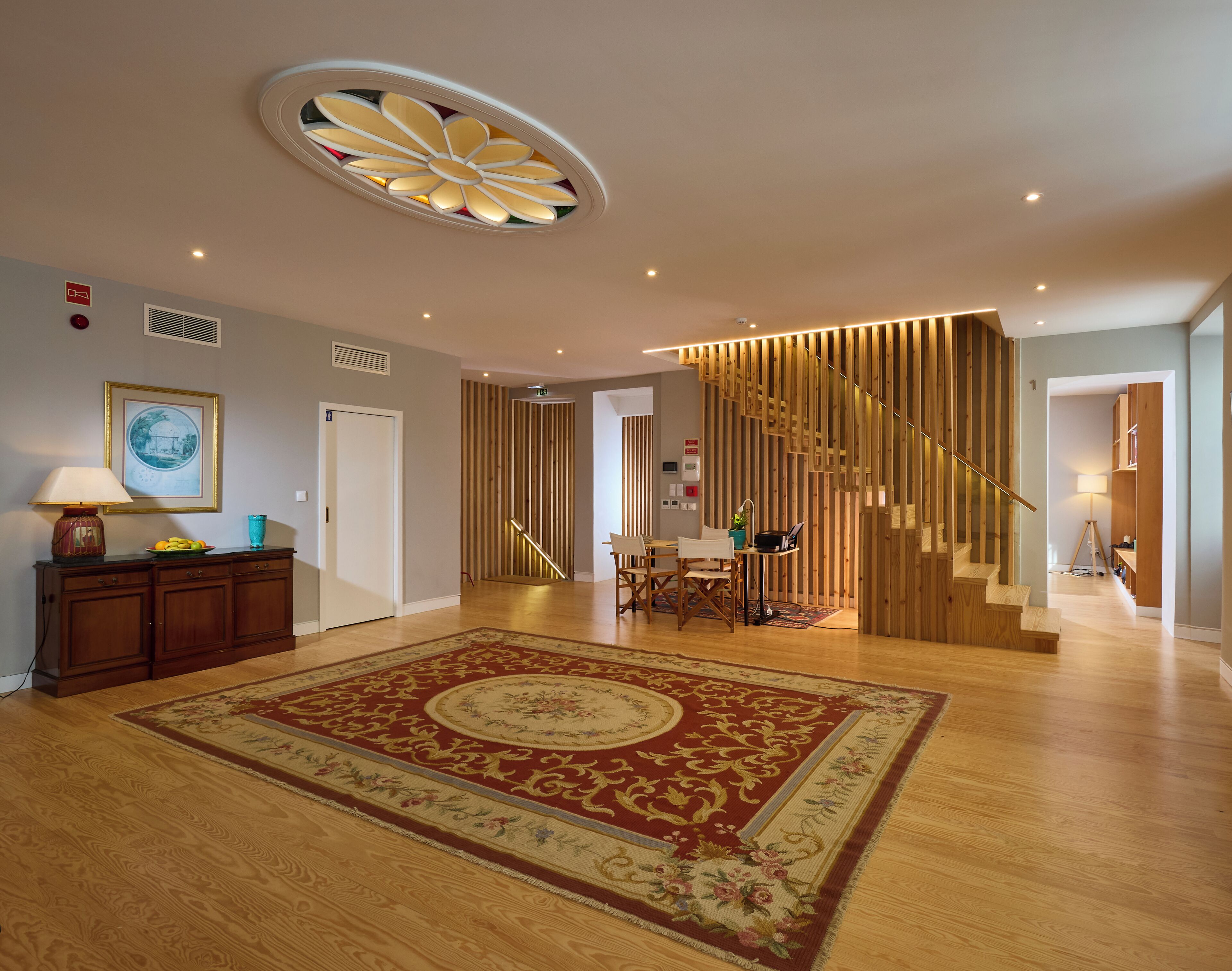 A spacious living area with a wooden floor, a large decorative rug, a staircase with wooden railings, and a ceiling with a circular light fixture.