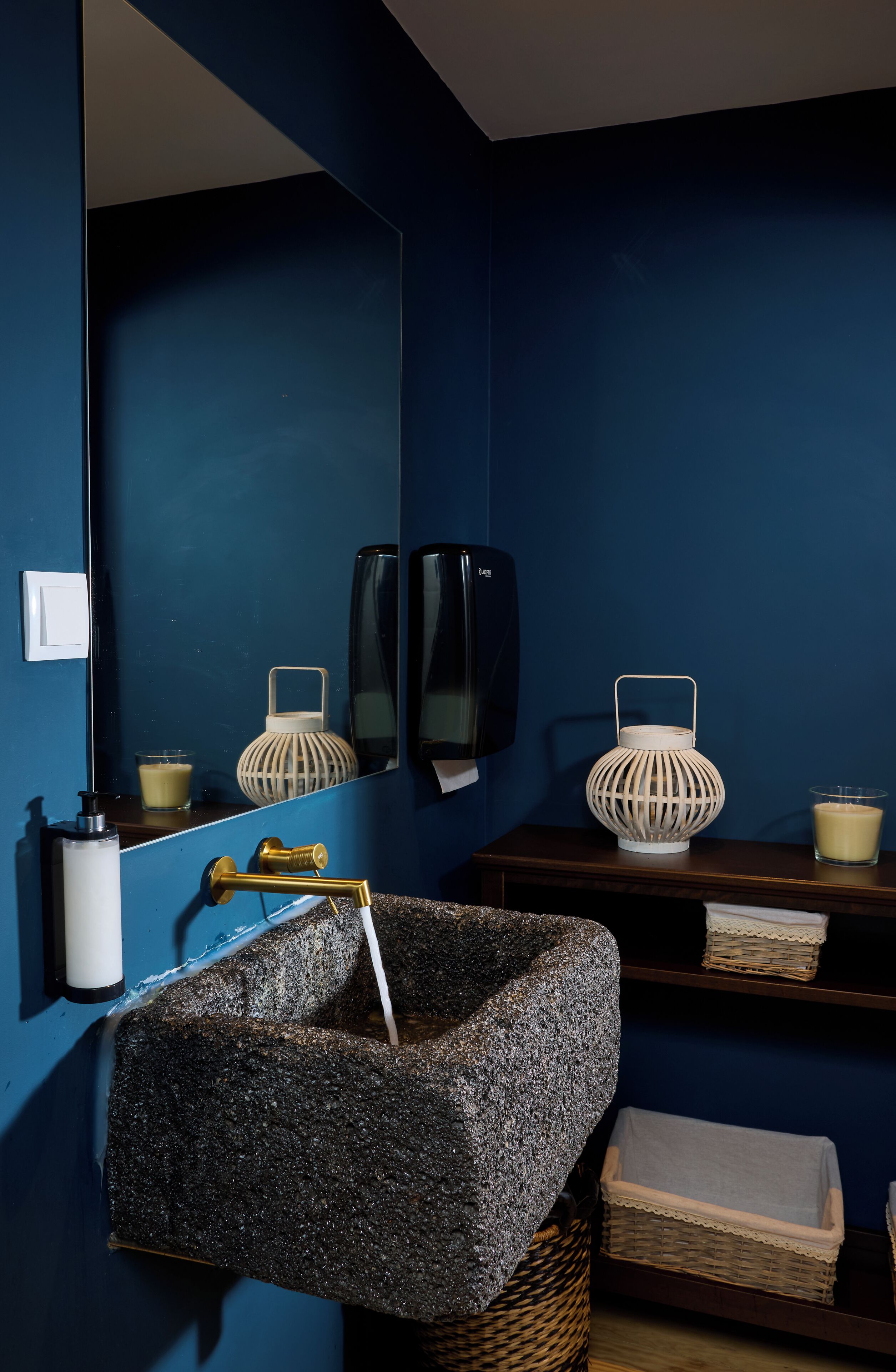 Casa de banho com parede azul, lavatório com cuba preta texturizada, espelho, dois castiçais brancos e prateleira de madeira com uma vela e um copo.