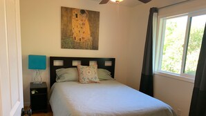 Een slaapkamer met een bed, een nachtkastje, een lamp, een plafondventilator, een raam met gordijnen, en een schilderij aan de muur.