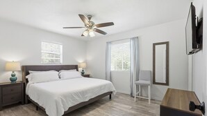 Ein Schlafzimmer mit einem Bett, Nachttischen, einem Deckenventilator, einem Sessel, einem Spiegel und einem an der Wand montierten Fernseher.