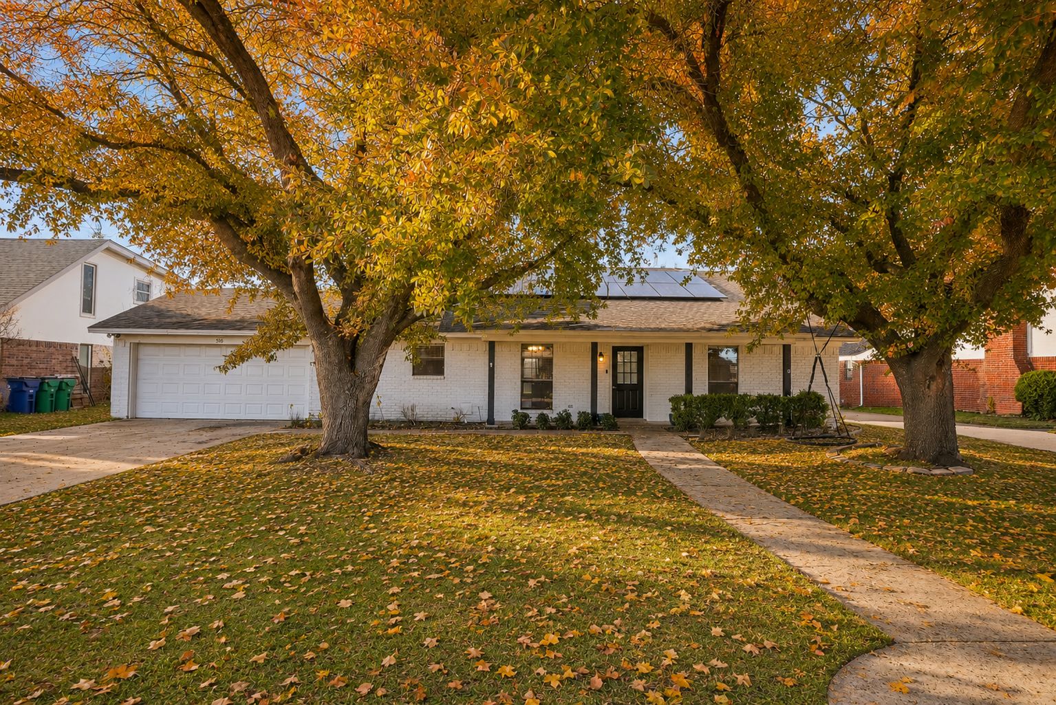 A residential house with a garage, a lawn with fallen leaves, and a tree in front.