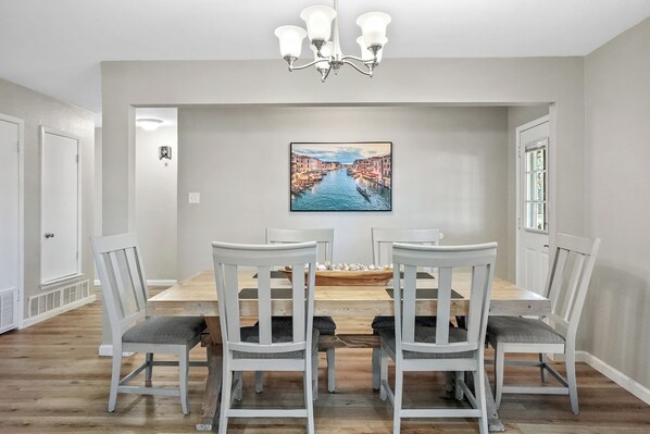 A dining room with a wooden table and white chairs, a chandelier, and a painting of a canal.
