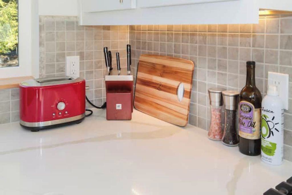 A kitchen counter with a red toaster, a wooden cutting board, a knife block, salt and pepper shakers, a bottle of olive oil, and a bottle of dish soap.