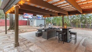 An outdoor patio with a covered seating area, stone flooring, and a TV screen displaying a sports event.