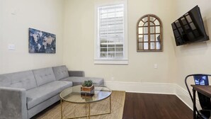 A living room with a grey sofa, a glass coffee table with a plant, a large window with blinds, and a mounted TV. - Cute centrally located 1bed 1bath walk to French Q, Bourbon St & Jazz venues  (New Orleans)