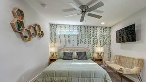 A bedroom with a bed, a ceiling fan, a window with hanging plants, and a wall-mounted TV. - The Southtown Hummingbird Haus (San Antonio)