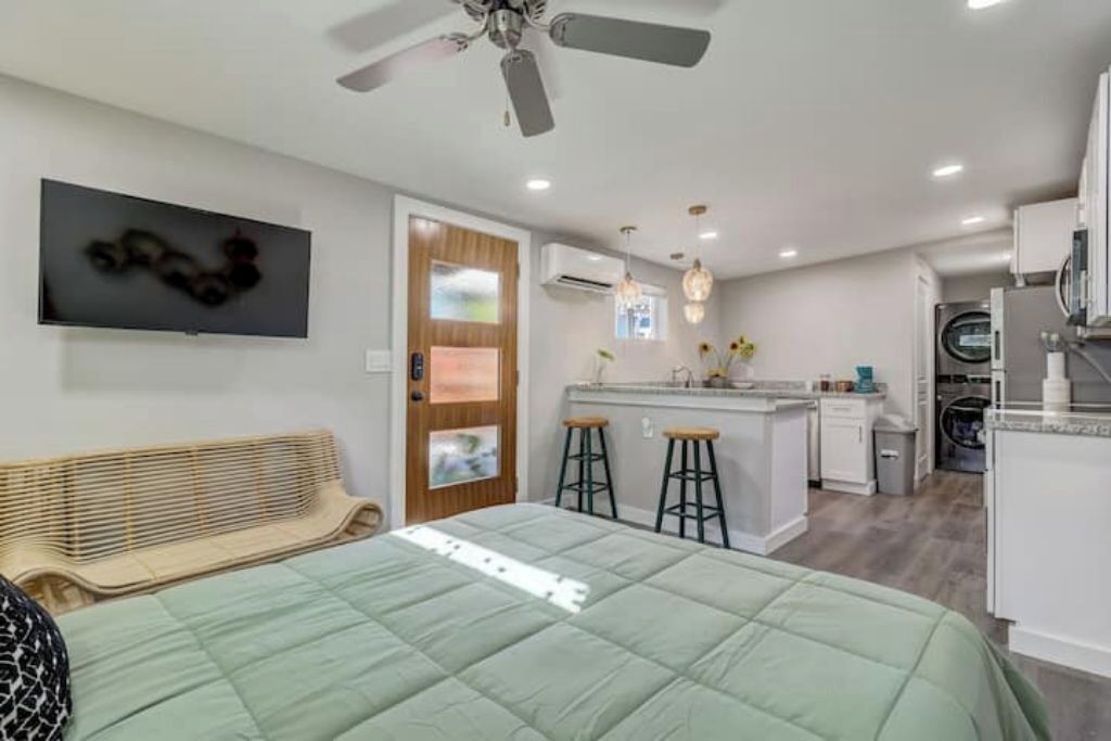 Quarto compacto com cama, ventilador de teto e TV na parede.