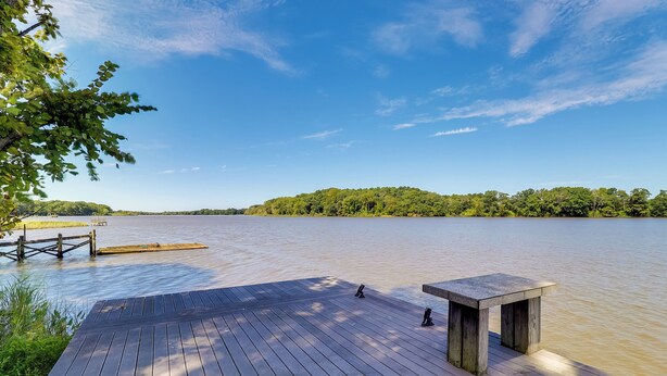 Eine Holzterrasse mit Tisch und Bänken bietet Blick auf einen stillen See, umgeben von Grün und wolkenlosem Himmel.