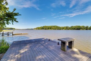 Um deck de madeira com mesa e bancos oferece vista para um lago calmo, cercado por vegetação e um céu claro.