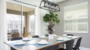 A dining area with a long table set for six, a potted plant, and a modern pendant light.