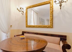 A dining area with a round wooden table, a cushioned bench, a gold-framed mirror, and a wall-mounted light fixture. - VILLA MIMOSA TAORMINA With Rooftop Sea View Terrace (Taormina)