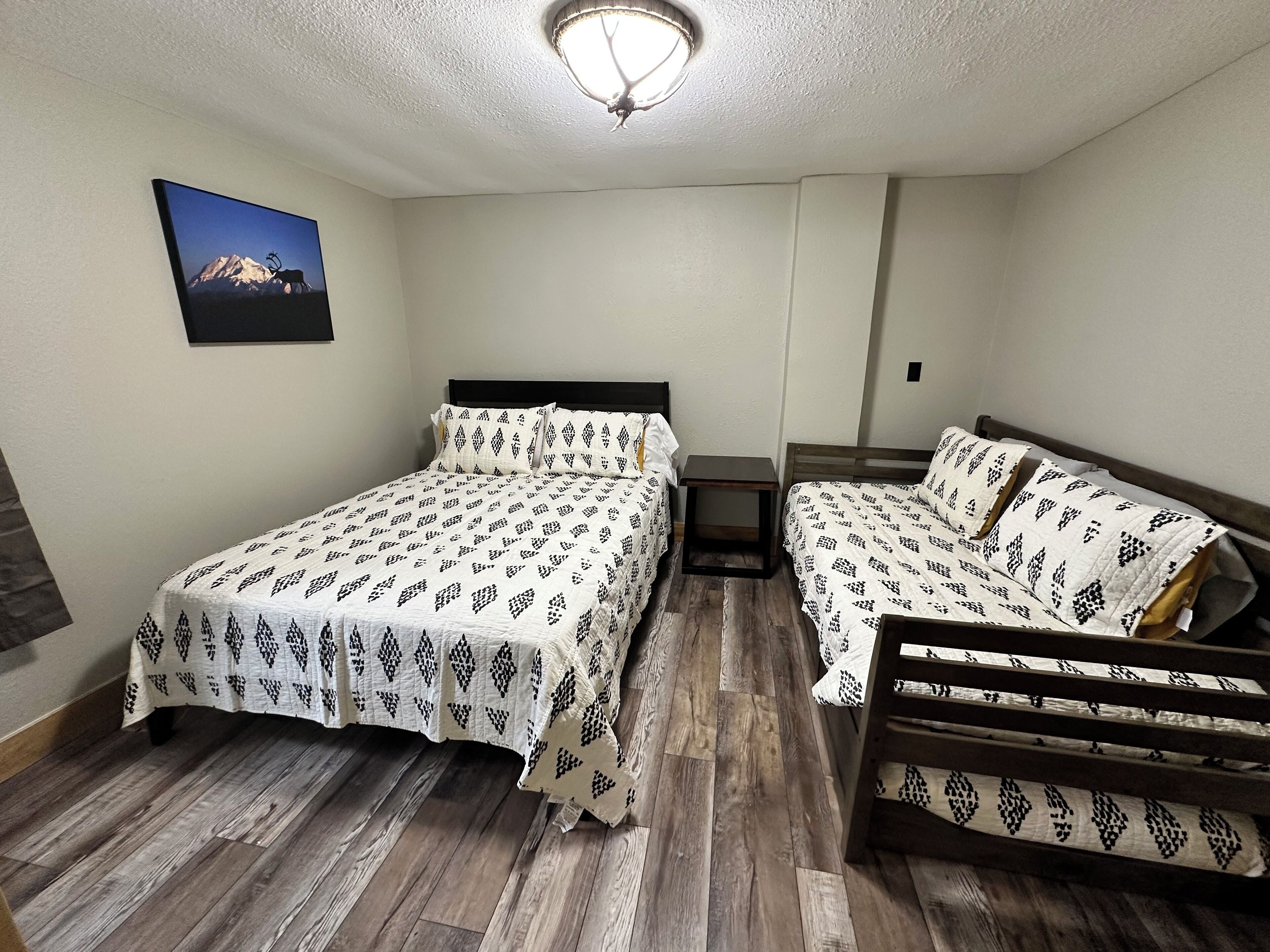 A bedroom with two beds, a wooden floor, a ceiling light, and a framed picture on the wall.