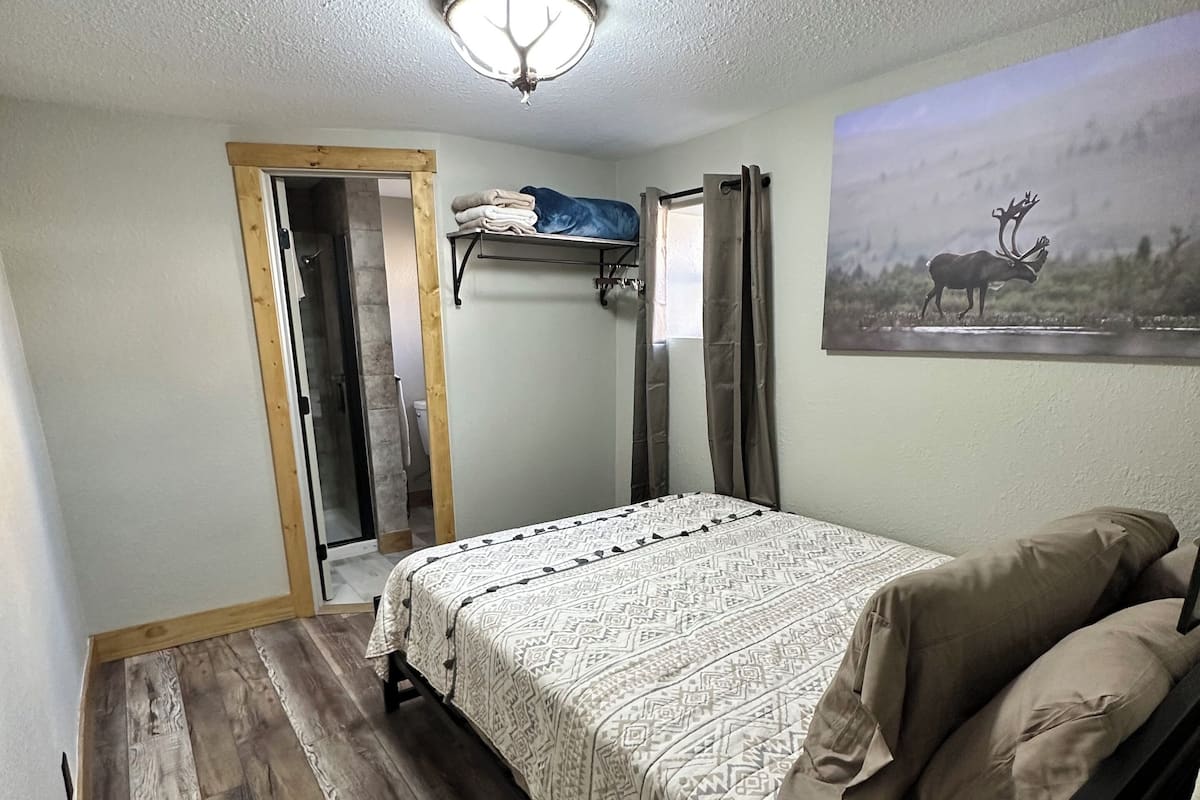 A bedroom with a bed, wooden flooring, a ceiling light, a door, and a wall-mounted shelf with folded clothes.