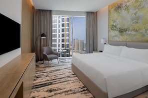 Habitación de hotel con una cama grande, un escritorio, una silla, una mesita y un ventanal con vistas a la ciudad.
