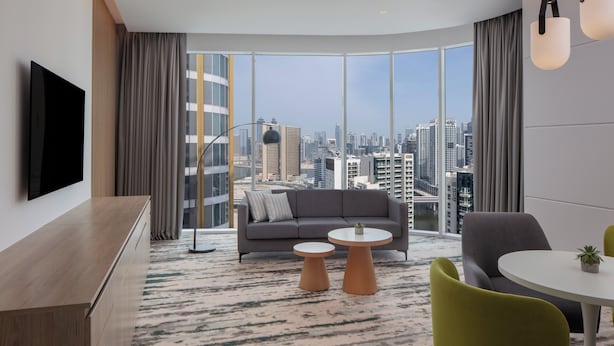 Habitación de hotel moderna con un ventanal grande que ofrece vistas a la ciudad, un sofá, una mesa de centro y un comedor con sillas.