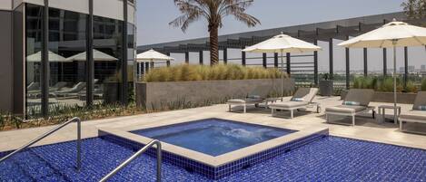 Una piscina sul tetto con piastrelle blu a mosaico, poltrona da relax, ombrelloni e una palma.
