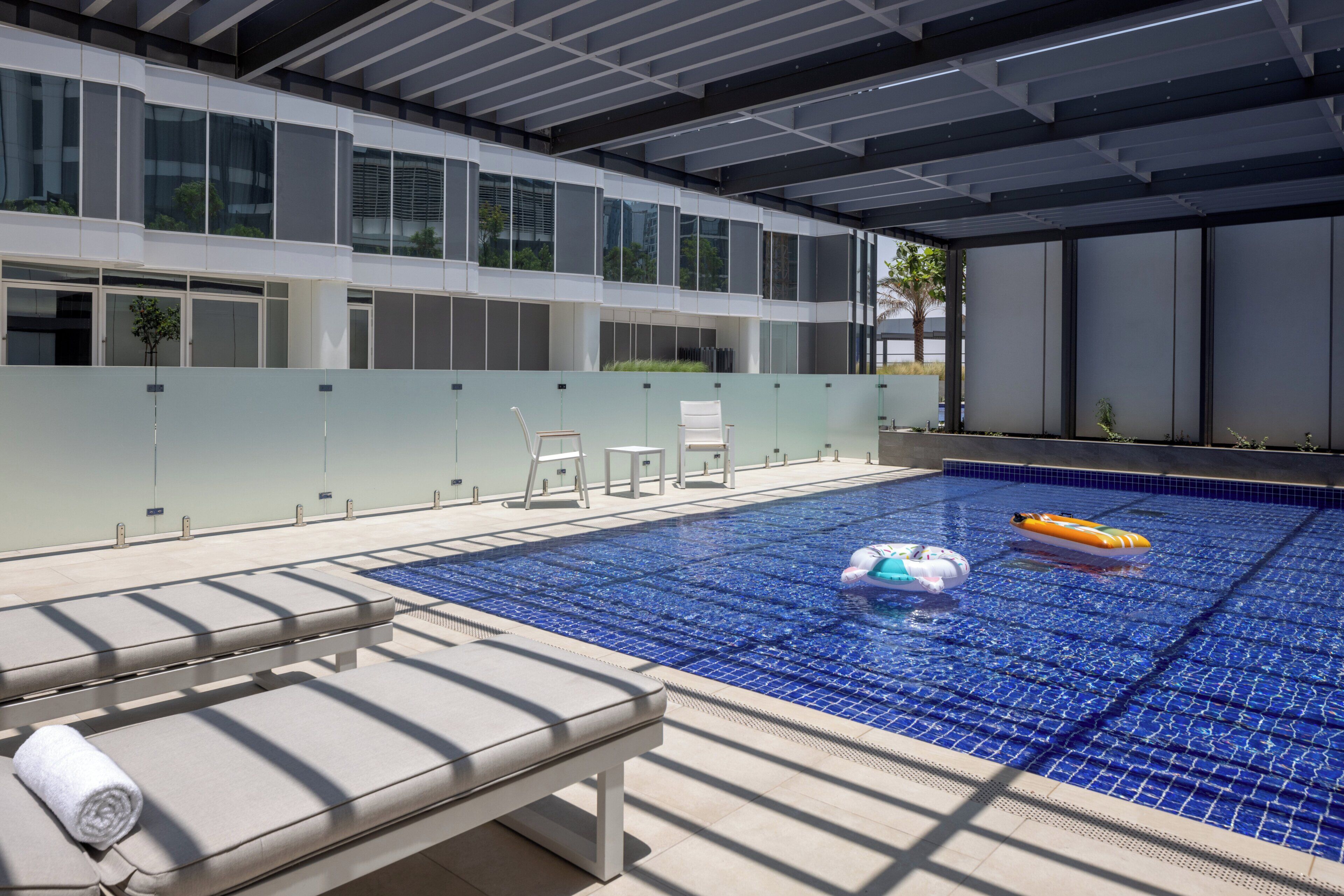 An indoor swimming pool with clear blue tiles, a lounge area with a bench and chairs, and a modern building facade with large windows.