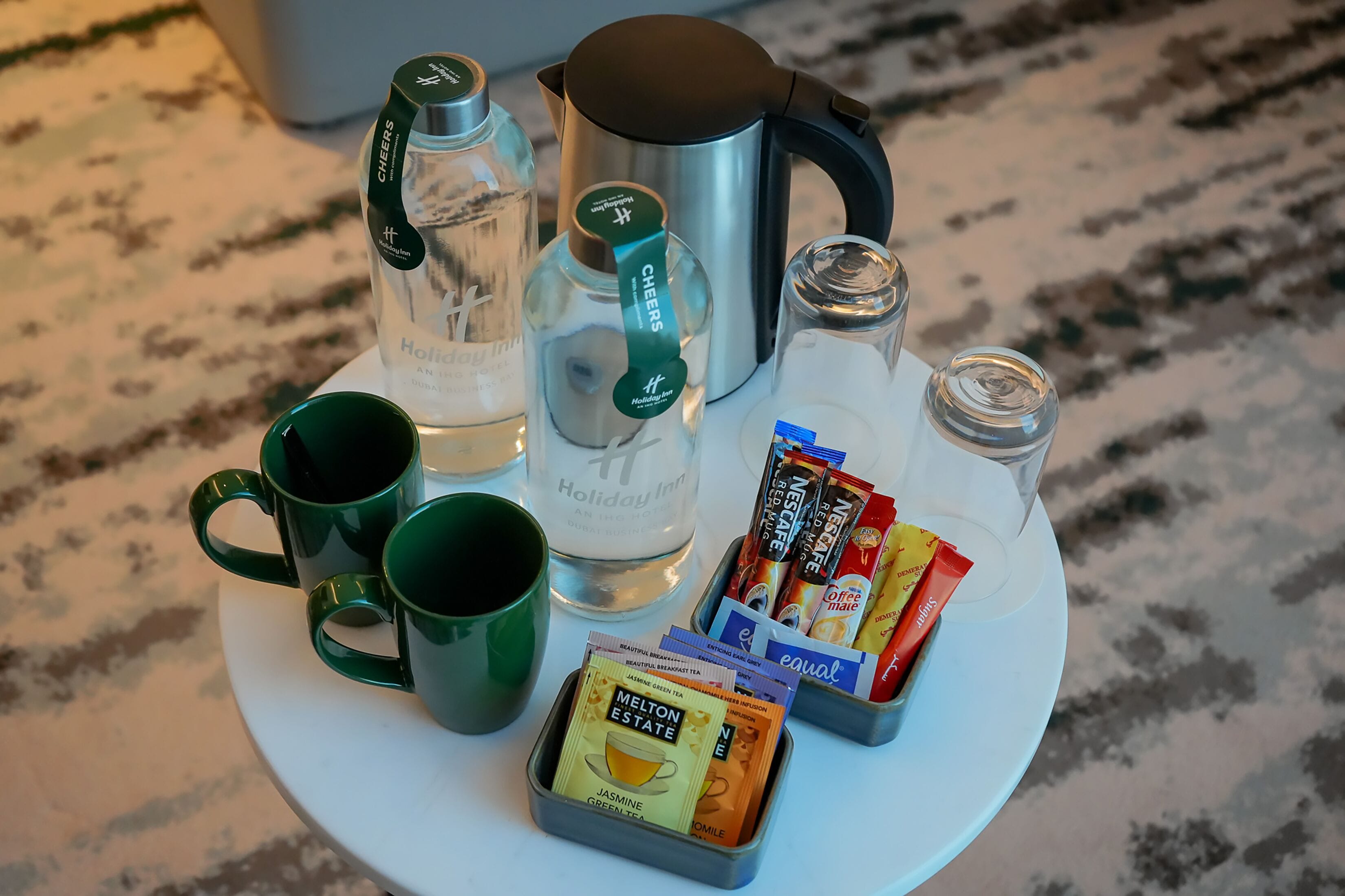 A table with a kettle, two mugs, a glass bottle, and several packets of tea and coffee.