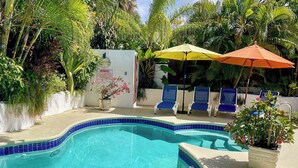 A swimming pool with blue-tiled edges, surrounded by lounge chairs and umbrellas, with lush greenery in the background. - Tropical Oasis with Pool close to beach - Beachcomber (Sunset Crest)