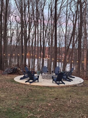 Un área de estar circular con sillas Adirondack alrededor de un hogar a leña de piedra, rodeada de árboles y un cuerpo de agua al fondo.