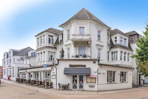 Ein weißes Gebäude mit Hauseingang an der Ecke, einem Balkon mit Statue sowie einer Ladeneinfahrt mit Außenbestuhlung.