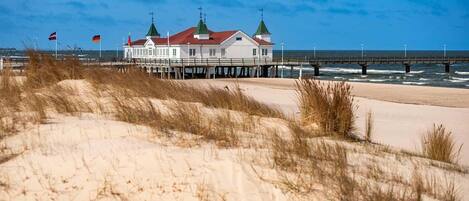 Ein Küstenhaus mit rotem Dach, weißen Wänden und grünen Türmchen, gelegen in der Nähe eines Sandstrandes und eines Piers.