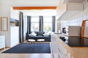 Cuisine moderne équipée d’un four à micro-ondes, d’une cuisinière et d’un four encastrés. Le salon comprend un canapé gris avec des coussins et une petite table.