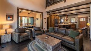 Un salón amplio con un sofá grande, dos sillones y una mesa de centro de madera. Se ve una cocina al fondo con armarios de madera y un salpicadero de piedra.