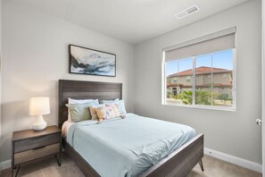 A bedroom with a bed, a nightstand, a lamp, a window with a view, and a ceiling vent.