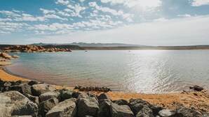 Una playa con grandes rocas al frente, agua tranquila y montañas en el fondo bajo un cielo parcialmente nublado.
