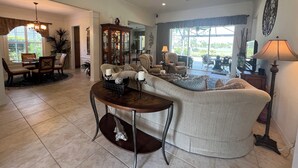 A living room with a curved sofa, a dining table with chairs, a chandelier, and a large window with curtains. - Luxurious golf home with pool in prestigious Colonial Country Club + Amenities (Fort Myers)