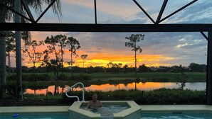 A person in a hot tub with a view of a sunset over a lake and trees. - Luxurious golf home with pool in prestigious Colonial Country Club + Amenities (Fort Myers)