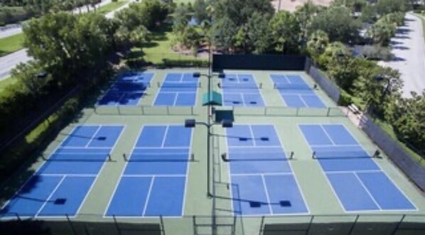 An outdoor tennis court complex with multiple courts, surrounded by trees and a fence.