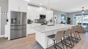 A modern kitchen with white cabinetry, stainless steel appliances, and a central island with bar stools. - OCEANLUX-Luxury+Gulf_Access+Heated_Pool+Kayaks+Golf+Chefs_Kitchen+Games (Cape Coral)