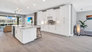 A modern kitchen with white cabinetry, a central island, and a large window overlooking a balcony. - OCEANLUX-Luxury+Gulf_Access+Heated_Pool+Kayaks+Golf+Chefs_Kitchen+Games (Cape Coral)