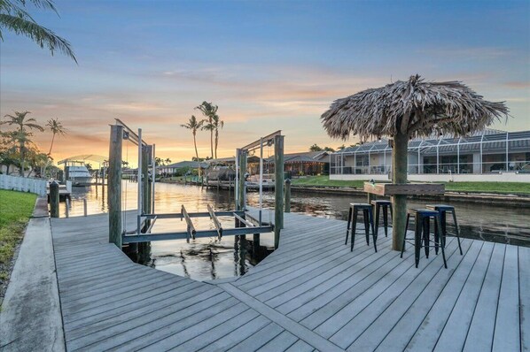 Un muelle de madera con un toldo de paja, una zona de bar y vistas a una vía fluvial con edificios al fondo.