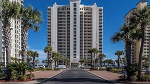 Un grand hôtel, avec une entrée centrale, entouré de palmiers et bordé de voitures garées.