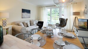 A well-lit living room with a sofa, a dining table set for two, and a television. - Haus Shelly - 3-zimmer Haus für 5 Personen: Terrasse, 2 Stellplätzen, Wlan. Haustier Erlaubt (Sylt-Ost)