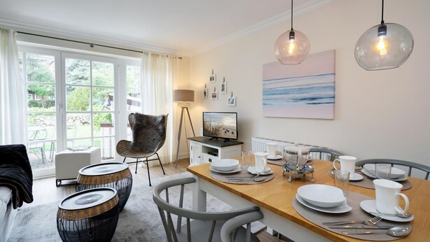 A dining area with a wooden table set for four, surrounded by chairs, and a large window providing a view of the outdoors. - Haus Shelly - 3-zimmer Haus für 5 Personen: Terrasse, 2 Stellplätzen, Wlan. Haustier Erlaubt (Sylt-Ost)