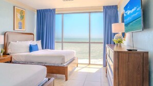 Habitación de hotel con cama, escritorio, televisor y ventana grande con vista al océano.