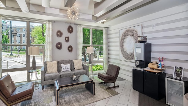 A modern living room with a sofa, coffee table, and chairs, large windows with curtains, a decorative wall piece, and a small refrigerator. - Uptown 4th Ward Luxury Apt Year-round Pool (Charlotte)