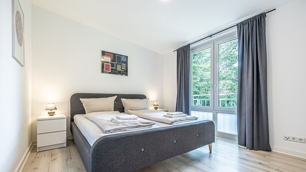 Ein modernes Schlafzimmer mit einem grauen Bett, weißen Bettwäsche, einem Nachttisch mit Lampe und einem großen Fenster mit Vorhängen.