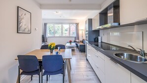 Eine moderne Küche mit einer großen Kochinsel, einem Essbereich mit zwei Stühlen und Blick ins Freie durch große Fenster.